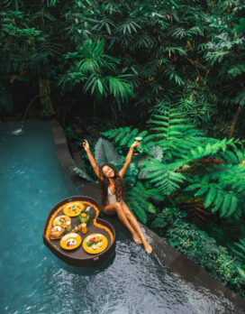 Happy girl relaxing and eating floating breakfast in jungle pool on luxury villa in Bali. Valentines day or honeymoon surprise. Tropical travel lifestyle. Black rattan tray in heart shape.
